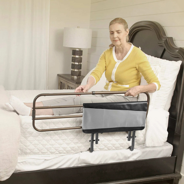 A woman in a yellow cardigan sits on the bed’s edge, holding the Signature Life Sleep Safe Extendable Bed Rail with an attached storage pouch for support and safety. See product specifications or warranty details for more information.