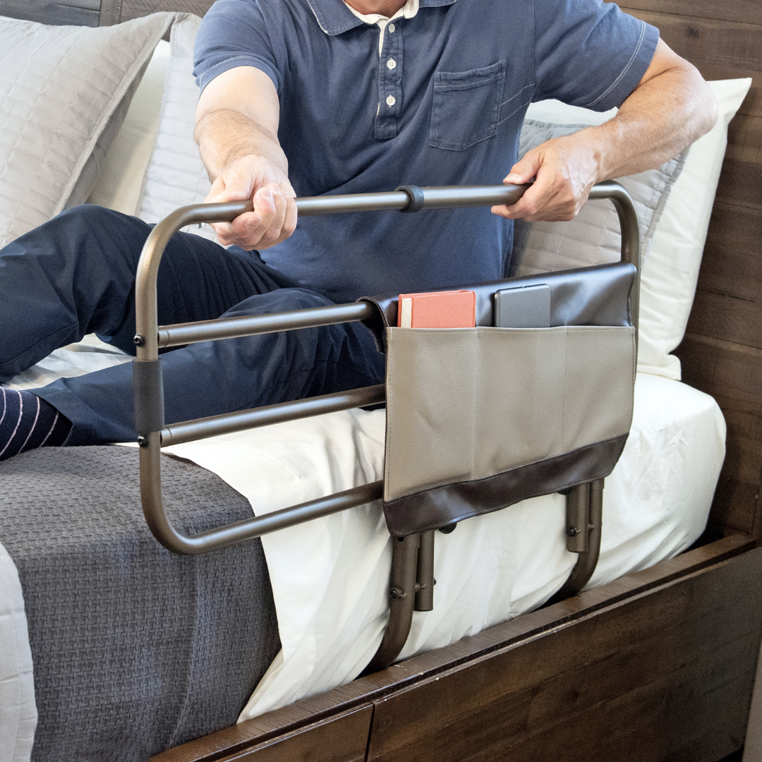 A person grips the Signature Life Sleep Safe Extendable Bed Rail, installed at the bedside for support. An attached pouch holds books, keeping personal items within easy reach. See Specifications for details.