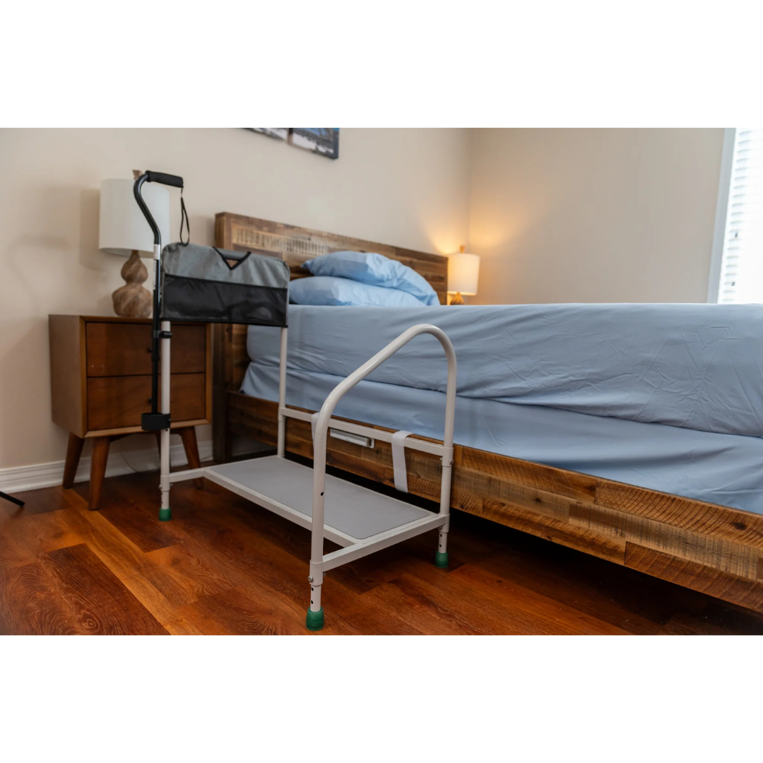 The Step2bed Deluxe Bedside Step-Up Platform with Accessory Kit, featuring a tray and pouch, is set up beside a wooden bed with blue sheets in a softly lit bedroom with hardwood floors and a nightstand.