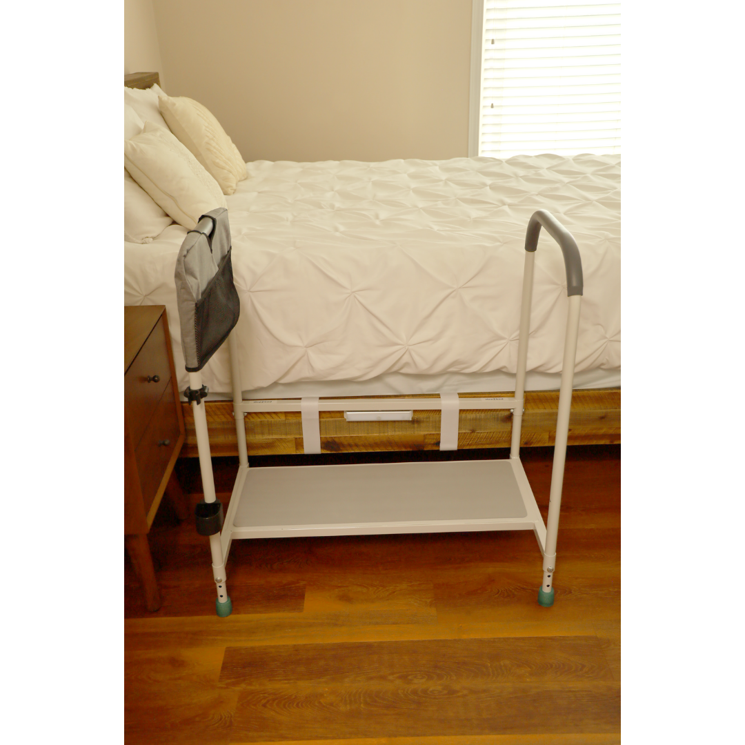 The Step2bed Deluxe Bedside Step-Up Platform with Accessory kit, featuring a bottom shelf, stands on the wooden floor by a nightstand beside a white bed safety rail with a support handle and side pouch, attached to the bed with a white comforter.