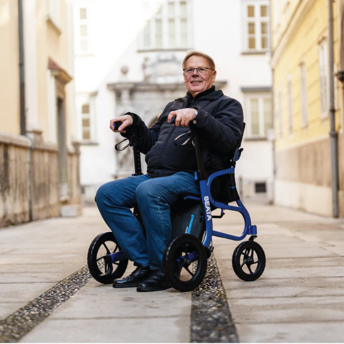 A person in a blue wheelchair, the Strongback Mobility SEATA Ergonomic Rollator, enjoys a day outside on a cobblestone street among buildings, smiling and wearing glasses, a black jacket, blue jeans, and black shoes.