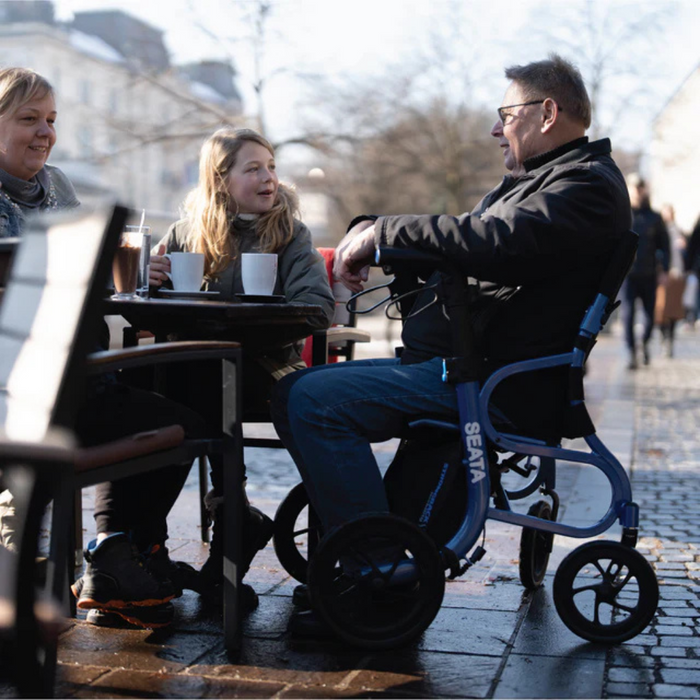 A man in a Strongback Mobility SEATA Ergonomic Rollator sits at an outdoor café table with a smiling woman and young girl. They enjoy drinks on a sunny day, surrounded by people walking in the background.