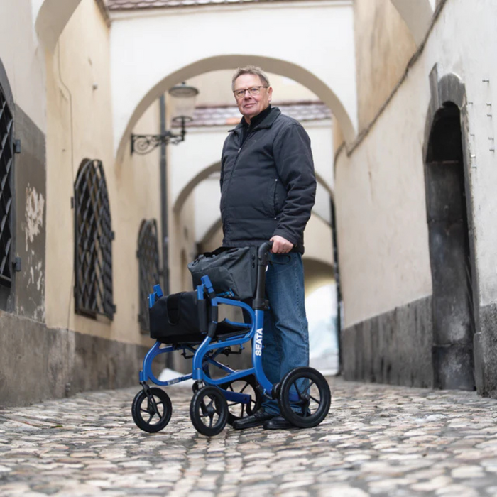 On a cobblestone street, a person in a dark jacket and glasses uses a Strongback Mobility SEATA Ergonomic Rollator. They navigate the narrow passage with arched ceilings and vintage lanterns, creating a historical ambiance that complements their lightweight rollator.