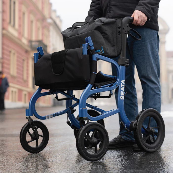 On a wet street, a person pushes the lightweight Strongback Mobility SEATA Ergonomic Rollator, featuring a black seat and storage pouch. Its ergonomic back support provides comfort as historic buildings loom in the cloudy background.