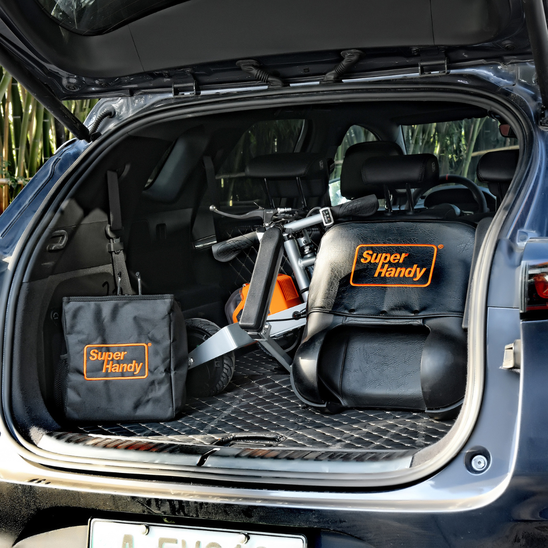 The car trunk holds a black Super Handy bag and a folded SuperHandy Passport Plus Portable Power Mobility Scooter - 11 Mile Range. Its compact, dual battery design allows for easy storage, and the spacious cargo area with protective mat is ideal for long-distance travel essentials.