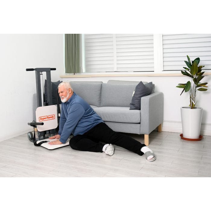 An elderly man is seated beside a gray couch, smiling and leaning on a stand labeled SuperHandy. The room features a white wall with a potted plant and gleaming wooden floors. His confidence is bolstered by the SuperHandy GoRise FS - Bariatric Electric Floor to Standing Lift Assistant.