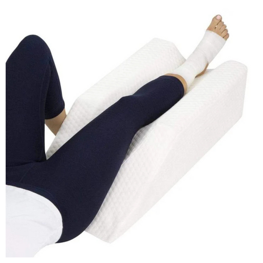 A person in dark leggings uses a Vive Health Knee Elevation Foam Pillow - Xtra-Comfort with Washable Cover, placing a bandaged leg on it for swelling reduction and surgery recovery support while lying down.