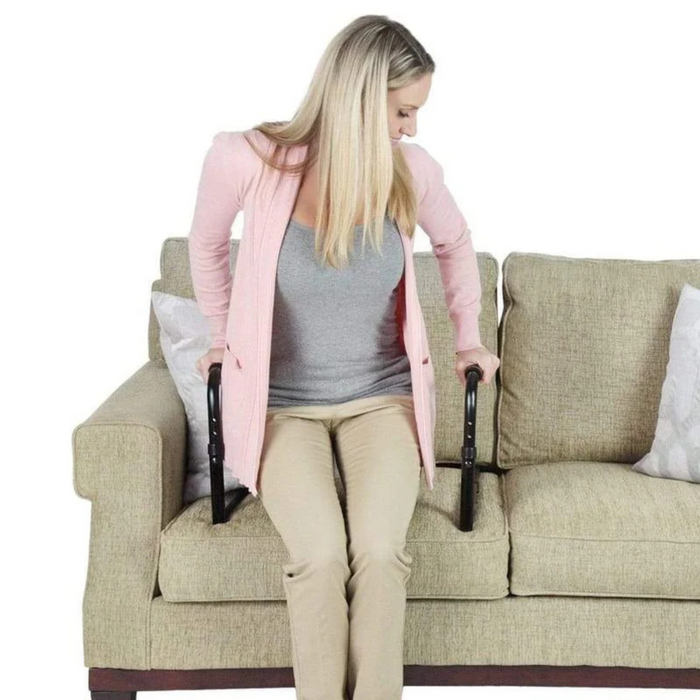 A woman wearing a pink cardigan and beige pants uses the Vive Health Couch Stand Assists black handles attached to a sofa, highlighting its tool-free assembly and adjustable aluminum frame for safe sitting and standing.